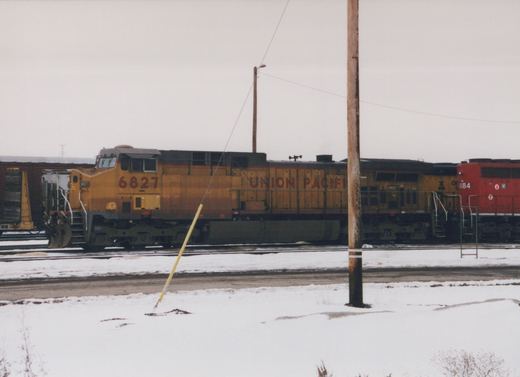 A yellow Union Pacific diesel locomotive numbered 6827, seen from the side on a yard track under a gray sky, with snow on the ground, a wooden utility pole in front and a red locomotive at the right edge.