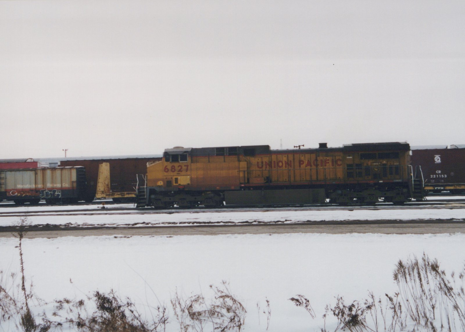 Side view of a yellow and gray Union Pacific diesel locomotive numbered 6827 standing on a yard track, with freight cars behind it and a snow-covered field with dry weeds in the foreground under an overcast sky.