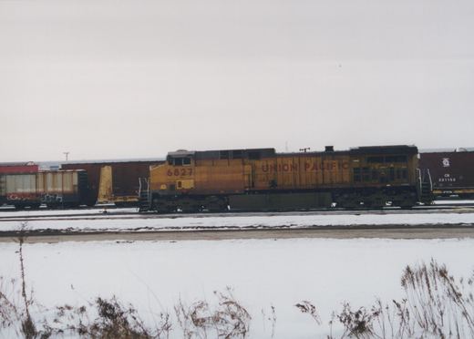 Side view of a yellow and gray Union Pacific diesel locomotive numbered 6827 standing on a yard track, with freight cars behind it and a snow-covered field with dry weeds in the foreground under an overcast sky.