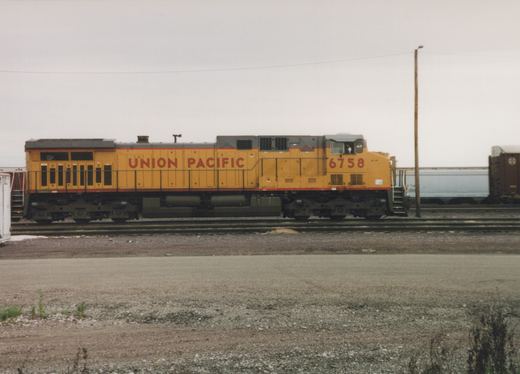 Side view of a yellow Union Pacific diesel locomotive numbered 6758 on a yard track, with freight cars behind it under an overcast sky.