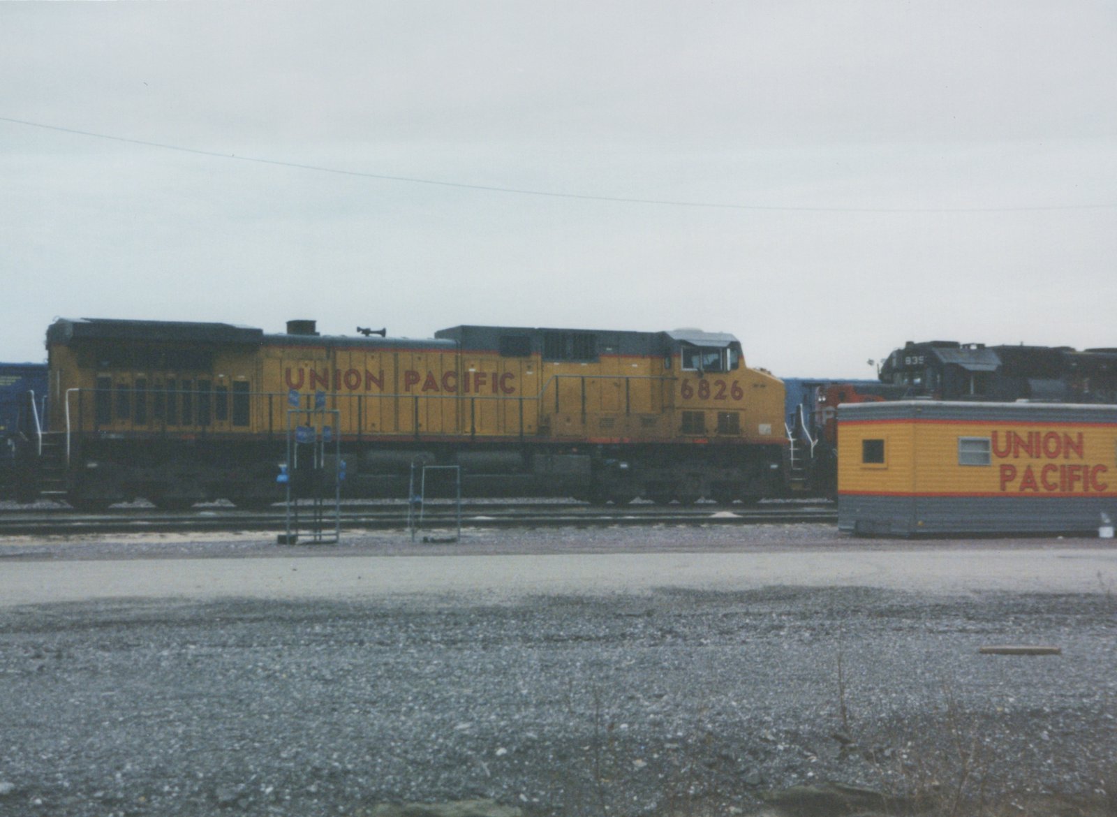 A yellow and gray Union Pacific diesel locomotive numbered 6826 seen from the side on a yard track under a gray sky, with a yellow Union Pacific yard building and a black locomotive at right.