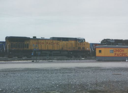 A yellow and gray Union Pacific diesel locomotive numbered 6826 seen from the side on a yard track under a gray sky, with a yellow Union Pacific yard building and a black locomotive at right.