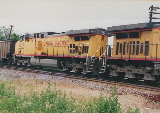 Two yellow Union Pacific diesel locomotives with red stripes and gray roofs, coupled rear to rear, pass on ballasted track under an overcast sky, with weeds in the foreground and a loaded coal car at the left.