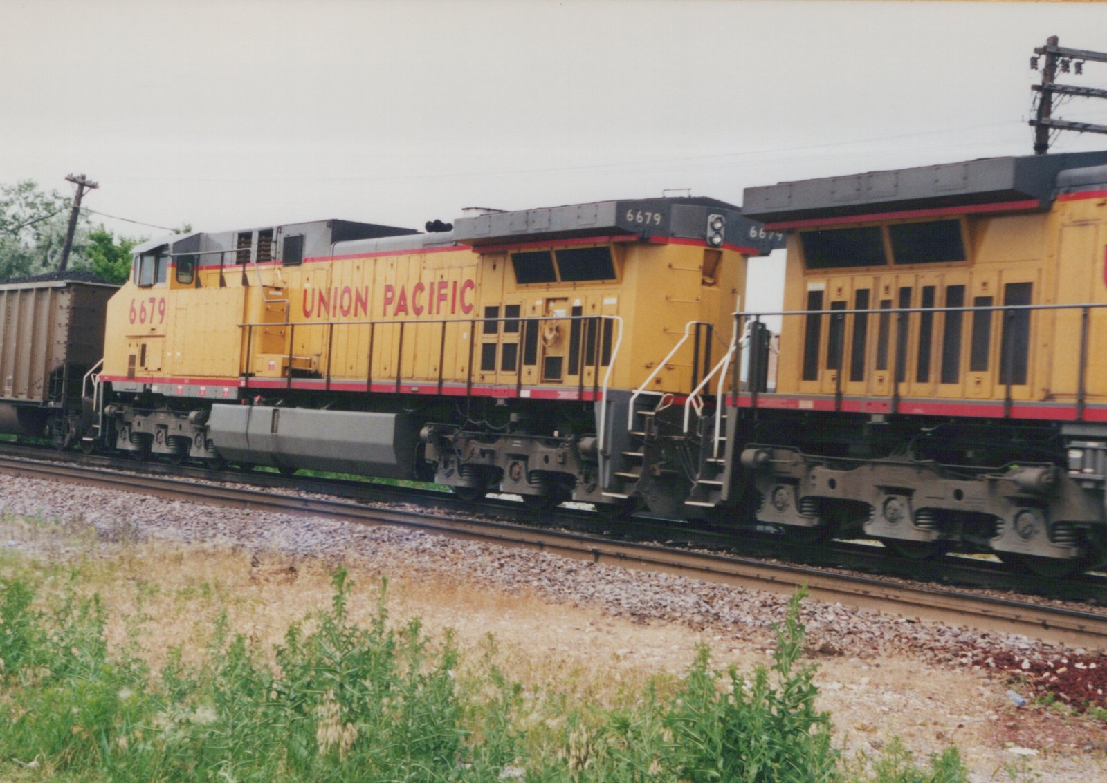 Two yellow Union Pacific diesel locomotives with red stripes and gray roofs, coupled rear to rear, pass on ballasted track under an overcast sky, with weeds in the foreground and a loaded coal car at the left.