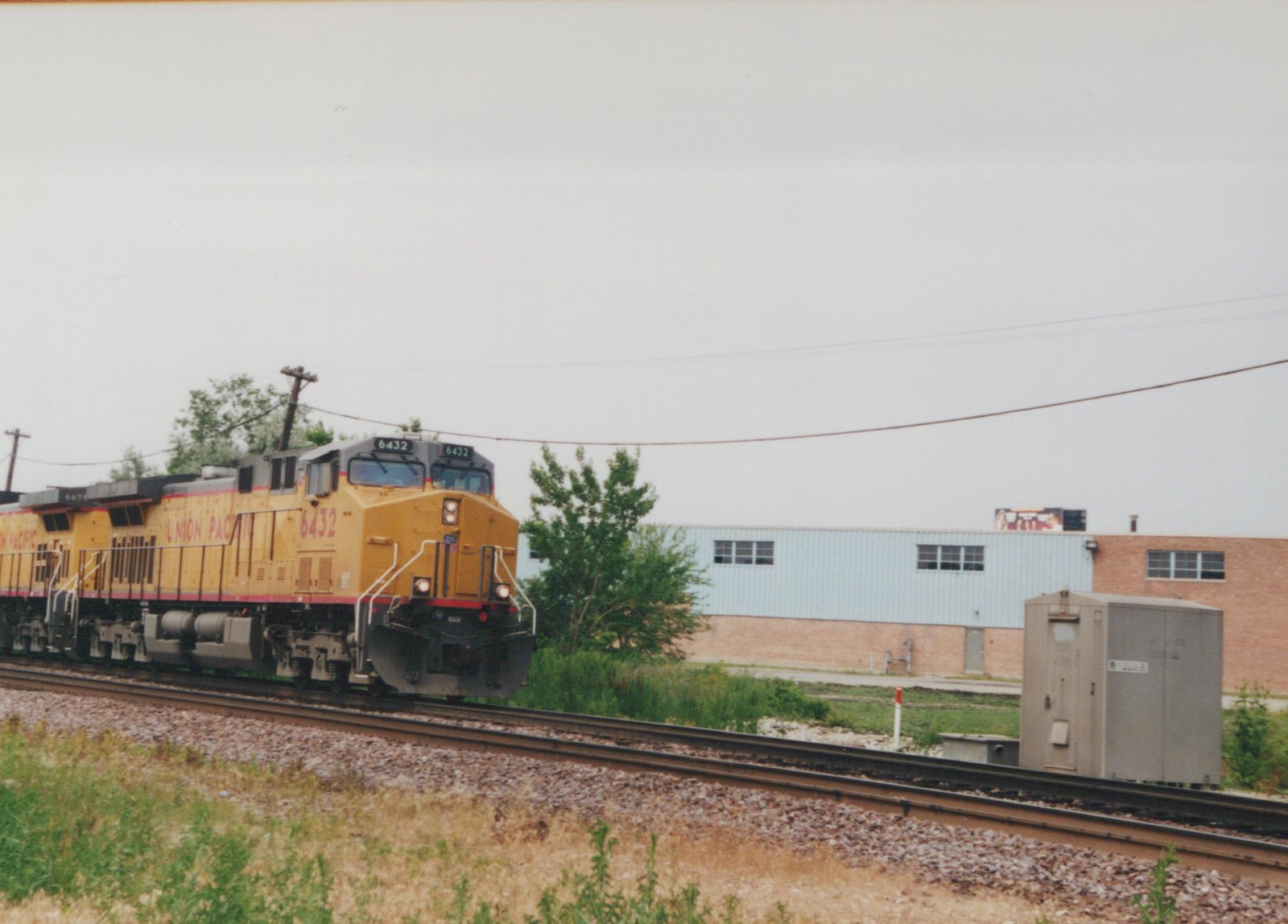 A yellow and gray Union Pacific diesel locomotive numbered 6432, with a second unit behind it, runs on a double-track line past a gray signal cabinet and a light-blue and brick industrial building.