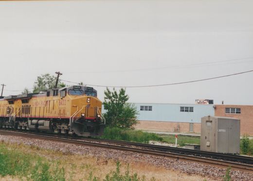 A yellow and gray Union Pacific diesel locomotive numbered 6432, with a second unit behind it, runs on a double-track line past a gray signal cabinet and a light-blue and brick industrial building.