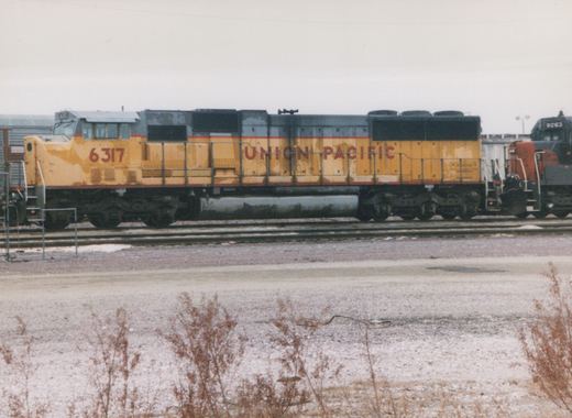 Side view of a faded yellow and gray Union Pacific diesel locomotive numbered 6317 on a yard track, a gray locomotive with a red nose numbered 9263 coupled behind it, under an overcast sky with dry weeds in the foreground.