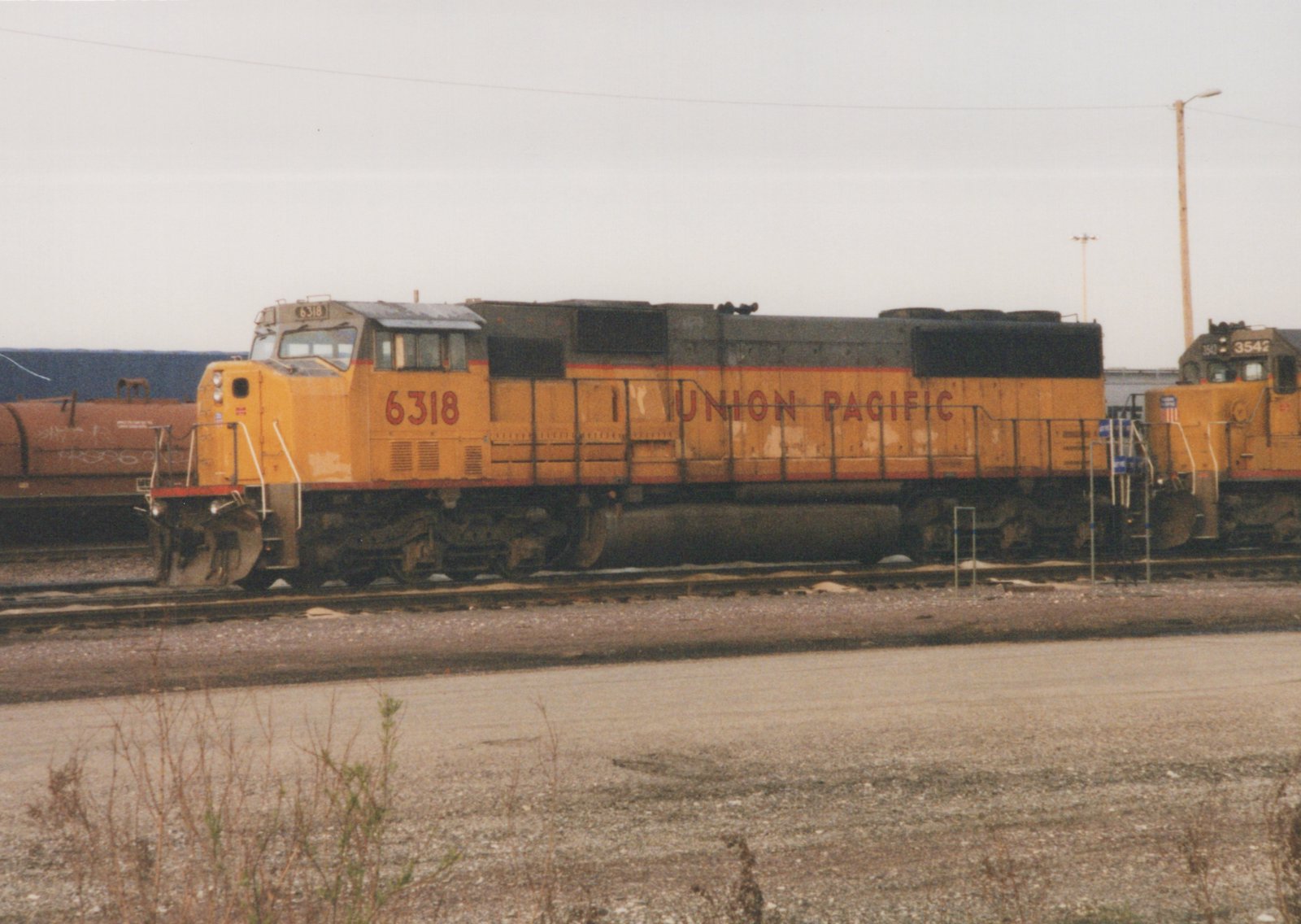 A weathered yellow and gray Union Pacific diesel locomotive numbered 6318 stands on a yard track with a second Union Pacific locomotive, numbered 3542, behind it, under an overcast sky.