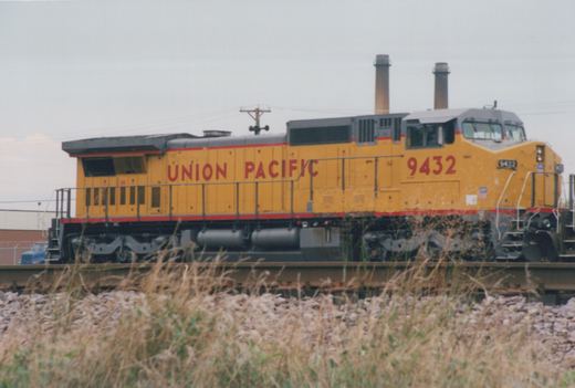 Side view of a yellow Union Pacific diesel locomotive numbered 9432 on a track, seen over tall dry grass, with two smokestacks and a utility pole under an overcast sky.