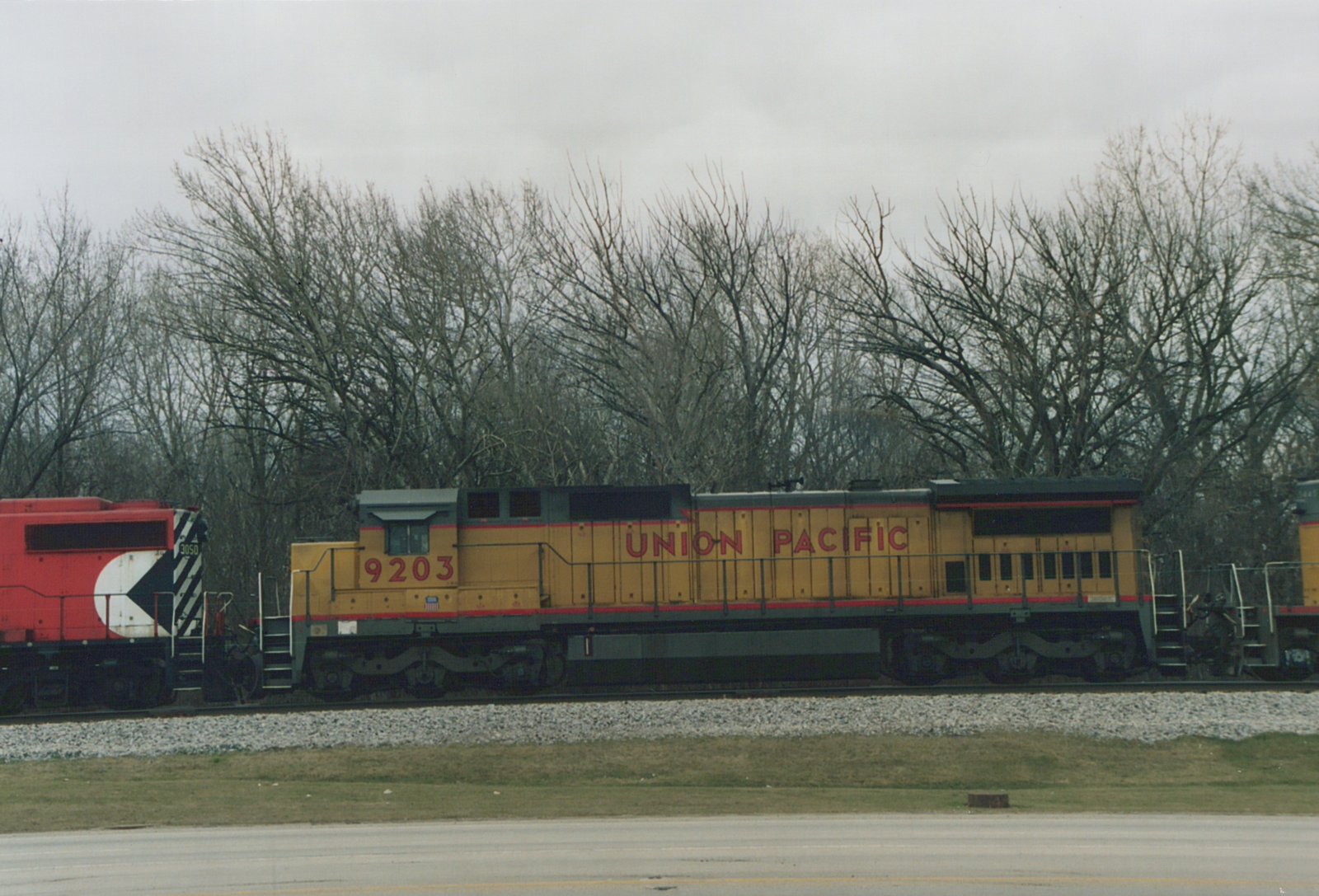 Side view of a yellow Union Pacific diesel locomotive numbered 9203 coupled between a red locomotive and another yellow unit, with bare trees and an overcast sky behind and a road in the foreground.