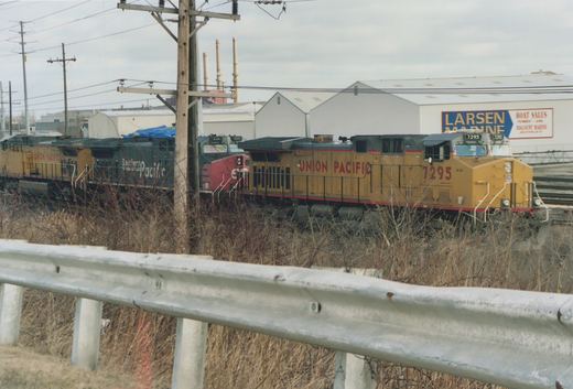 Three diesel locomotives, a yellow Union Pacific unit in front, then a gray and red Southern Pacific unit and a second yellow Union Pacific unit, seen behind a highway guardrail and dry brush, with warehouses and tall smokestacks under an overcast sky.