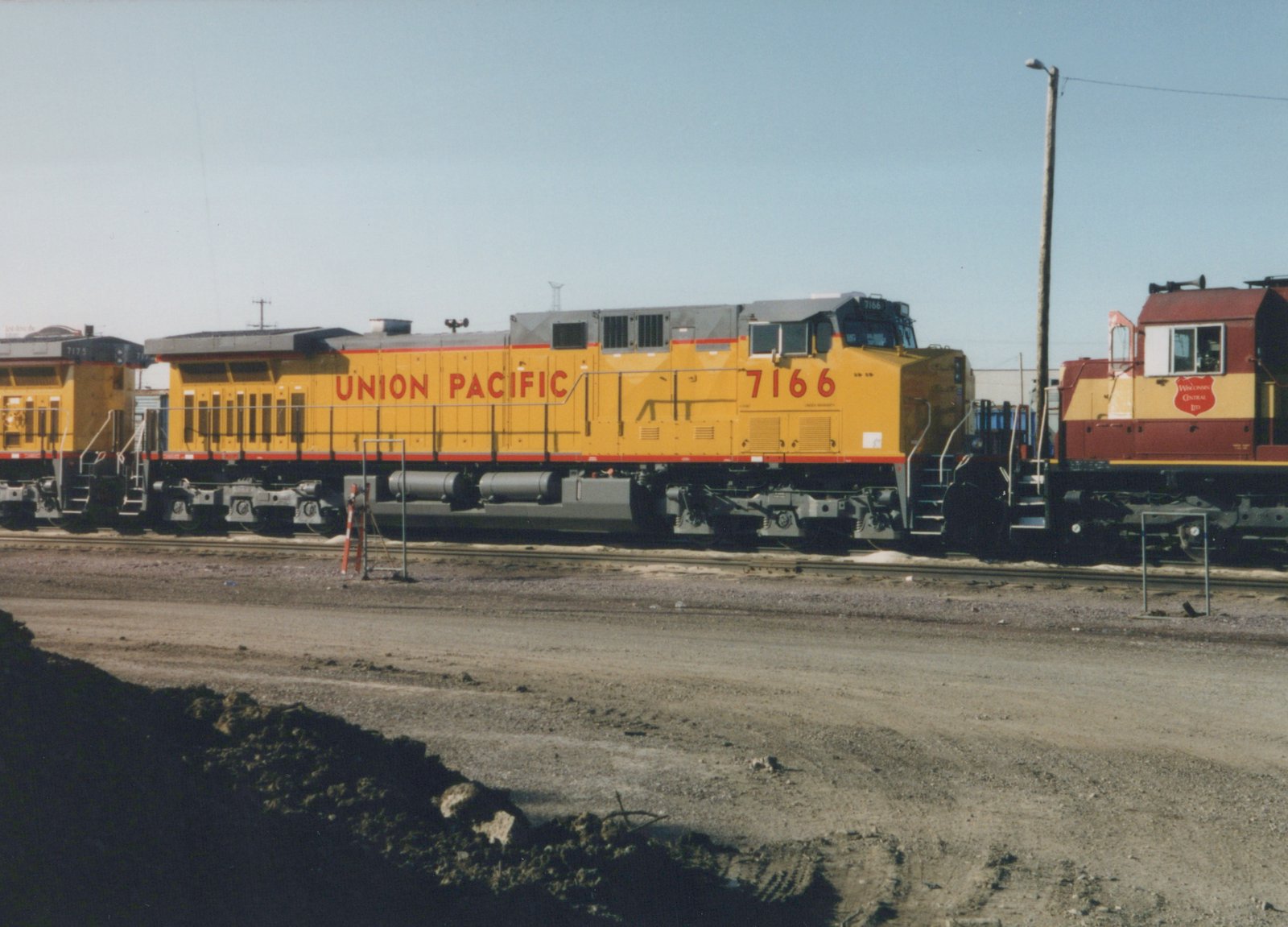 Side view of a yellow Union Pacific diesel locomotive numbered 7166, with another yellow unit behind it and the cab of a maroon and gold Wisconsin Central Ltd. locomotive ahead, on yard tracks under a clear sky with a dirt road and mound in the foreground.