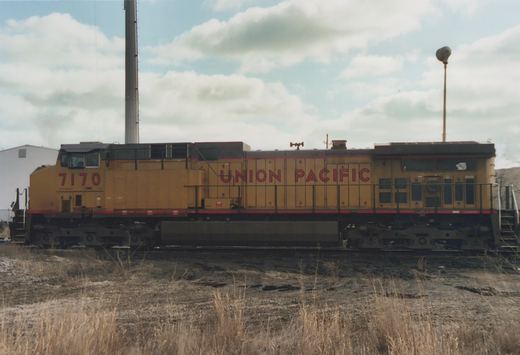Side view of a yellow Union Pacific six-axle diesel locomotive numbered 7170 on a track. A tall smokestack, a light pole and a white building are behind it, and dry grass is in the foreground.