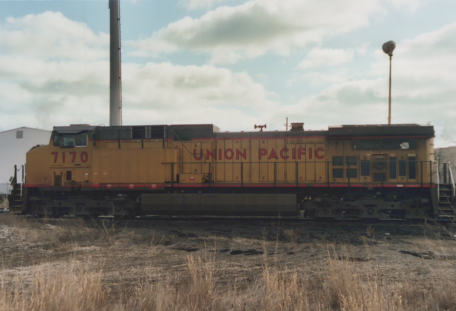 Side view of a yellow Union Pacific six-axle diesel locomotive numbered 7170 on a track. A tall smokestack, a light pole and a white building are behind it, and dry grass is in the foreground.