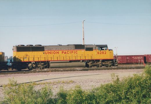 A yellow and gray Union Pacific diesel locomotive numbered 6282 is shown in profile next to a line of maroon Wisconsin Central boxcars under a clear blue sky.