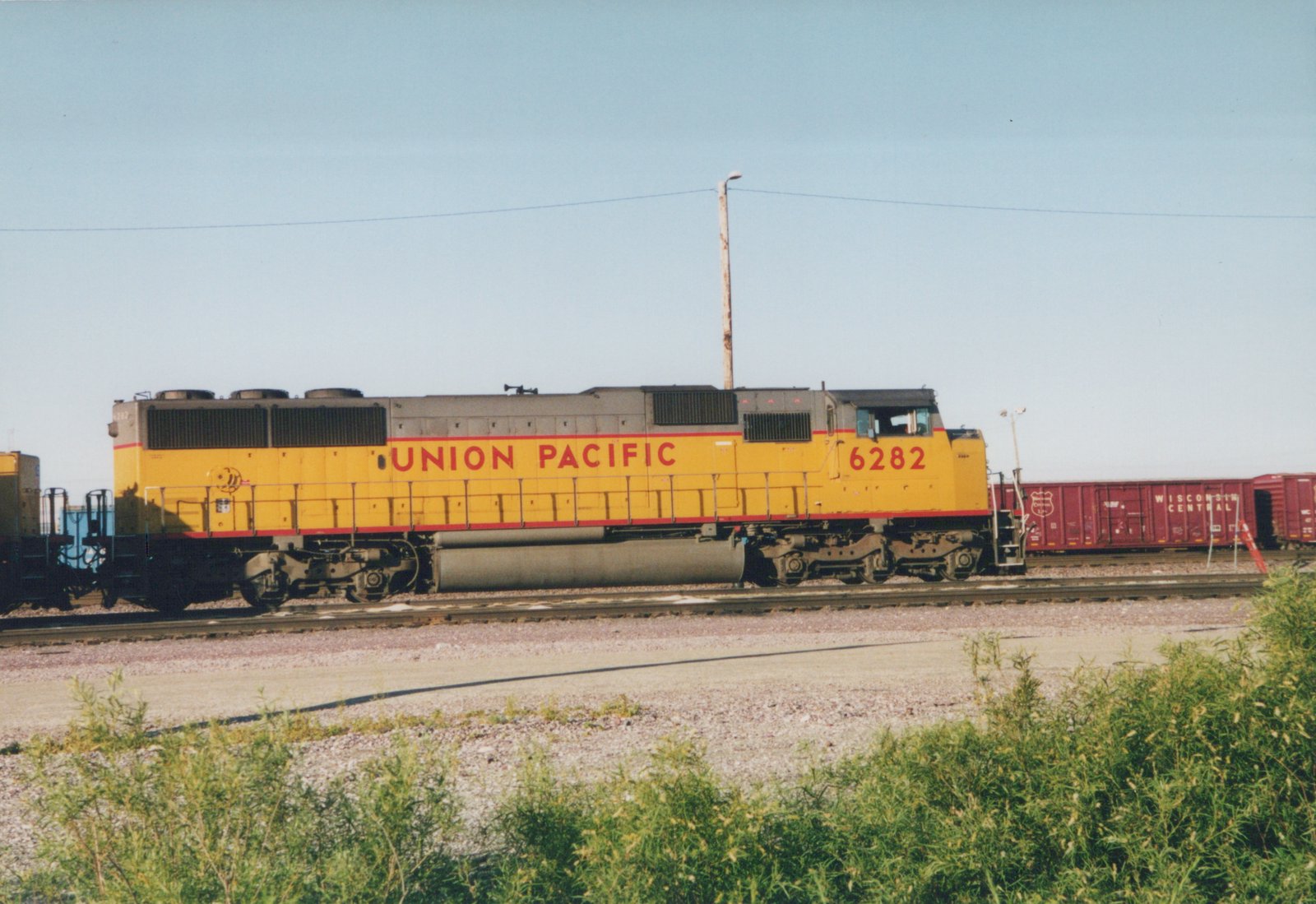 A yellow and gray Union Pacific diesel locomotive numbered 6282 is shown in profile next to a line of maroon Wisconsin Central boxcars under a clear blue sky.