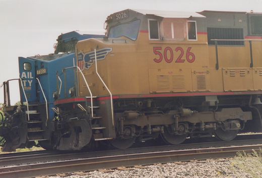 A yellow Union Pacific locomotive numbered 5026 is coupled to a blue RailTY locomotive numbered 7302, both on railroad tracks in a yard.
