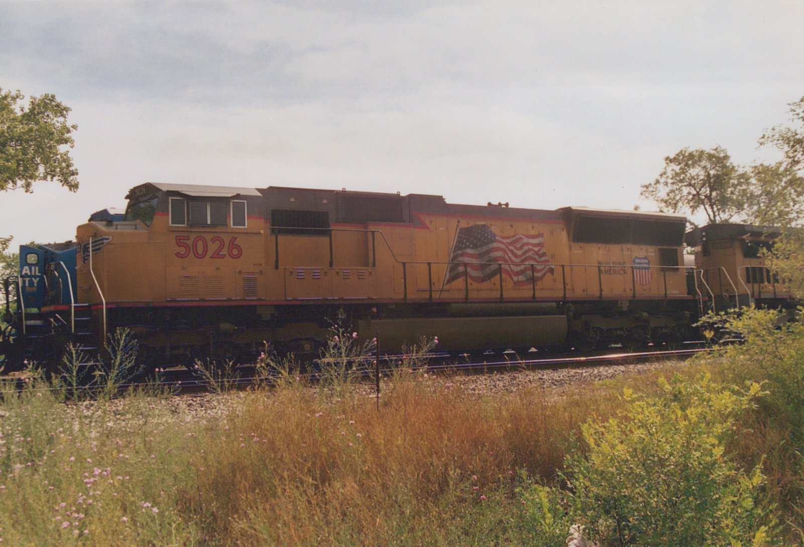 Side view of a yellow Union Pacific locomotive with the number 5026 and an American flag graphic, standing on railroad tracks beside grassy vegetation under a cloudy sky.
