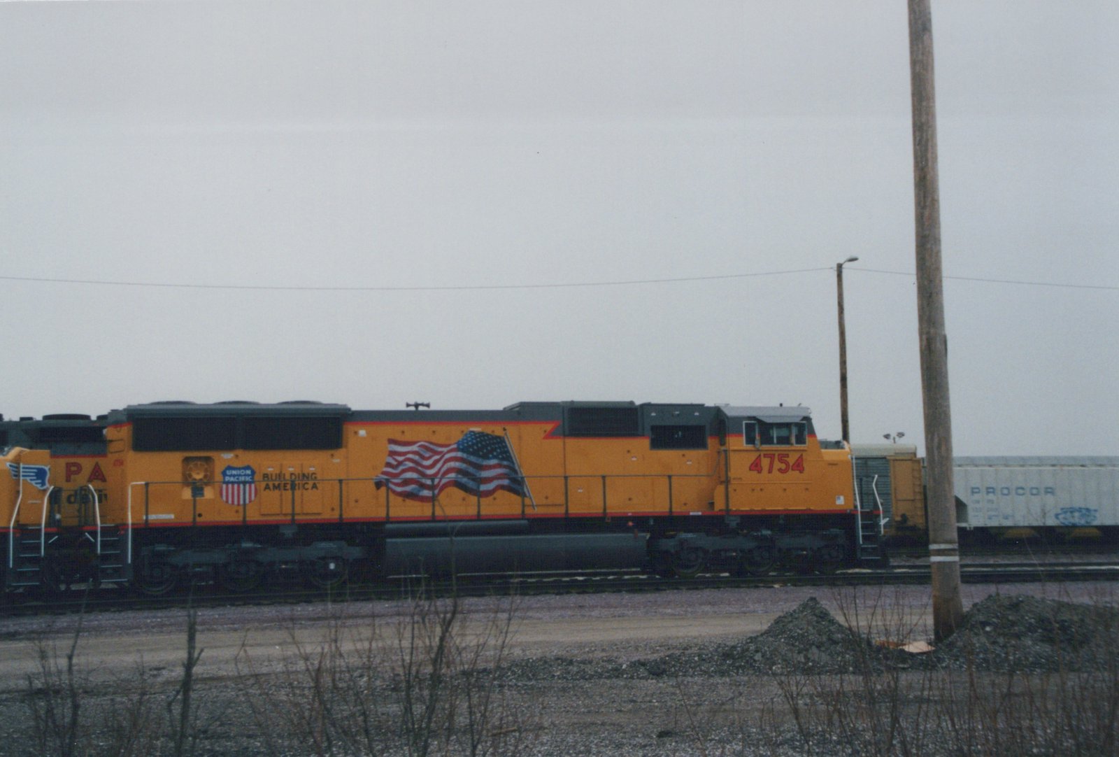 A yellow Union Pacific diesel locomotive numbered 4754 with a large American flag on its side sits on railroad tracks in a winter yard, with a white freight car labeled PROCOR in the background.