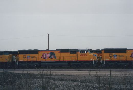 Two orange Union Pacific locomotives numbered 4753 and 37 sit on tracks in a yard under an overcast sky, with a large American flag painted on their sides.
