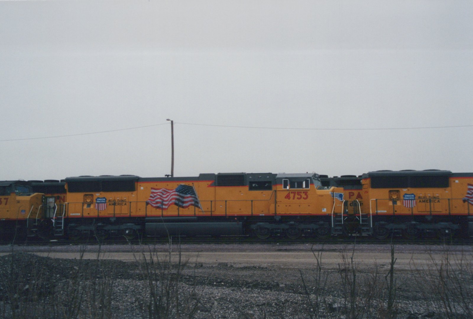 Two orange Union Pacific locomotives numbered 4753 and 37 sit on tracks in a yard under an overcast sky, with a large American flag painted on their sides.