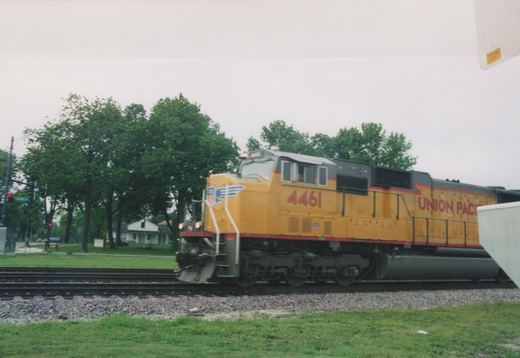 A yellow Union Pacific locomotive with the number 4461 is moving on train tracks beside a grassy area with trees and a grade crossing in the background.