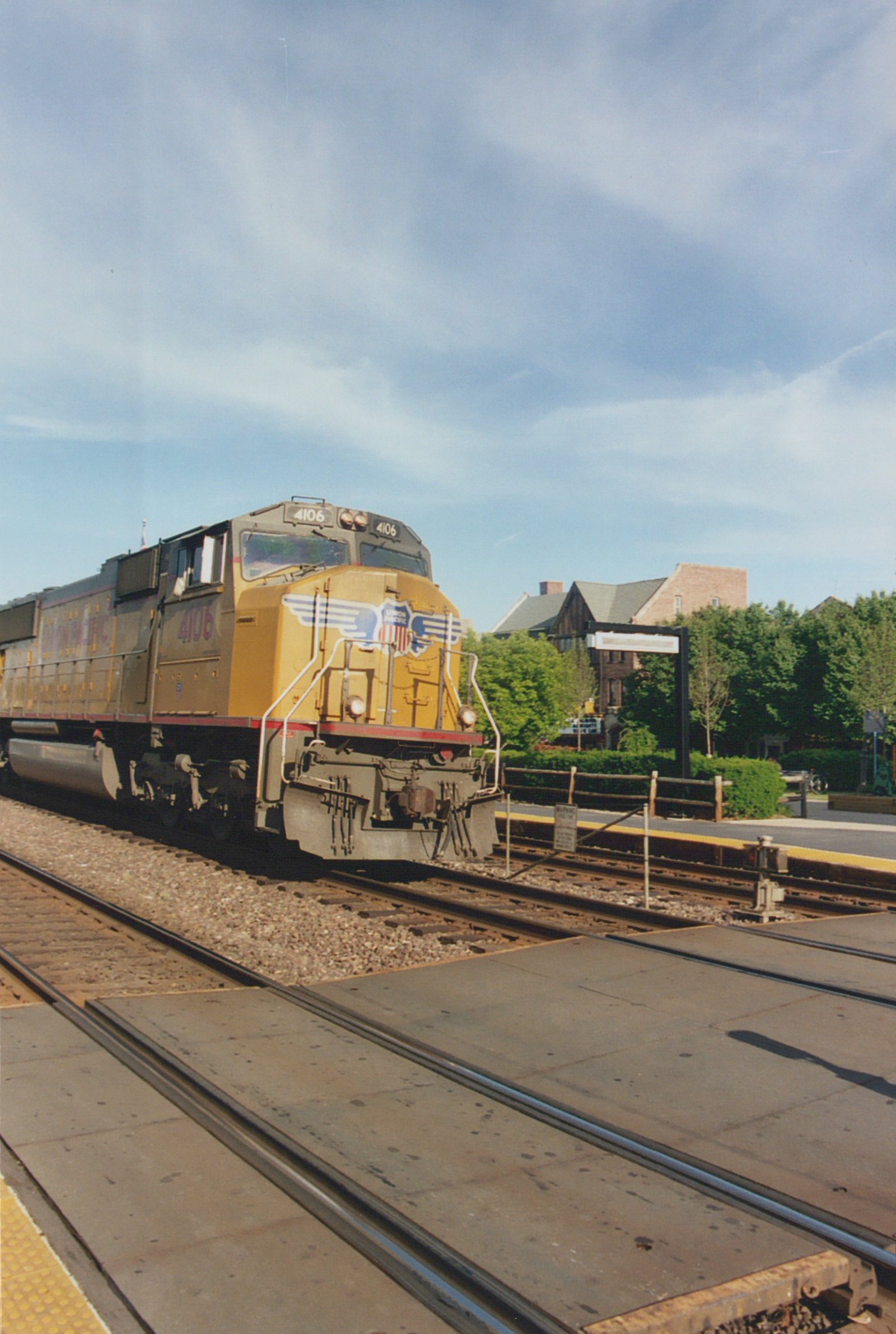 A yellow and gray Union Pacific locomotive numbered 4106 is stopped at a station platform with trees and buildings in the background under a blue sky.