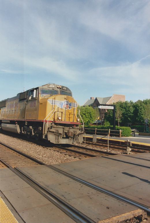 A yellow and gray Union Pacific locomotive numbered 4106 is stopped at a station platform with trees and buildings in the background under a blue sky.