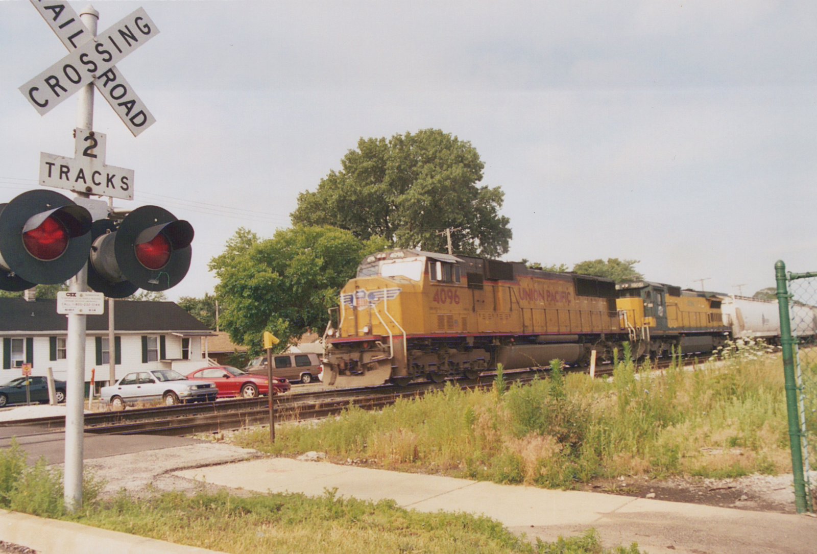 A yellow and black Union Pacific locomotive with the number 4096 is crossing a street at a railroad crossing with flashing red lights; several cars are parked nearby and a white house is visible in the background.