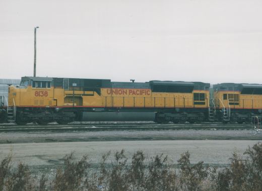 A yellow and gray Union Pacific locomotive with the number 8138 on its side is stopped on the tracks in a rail yard, with another locomotive visible behind it under a gray sky.