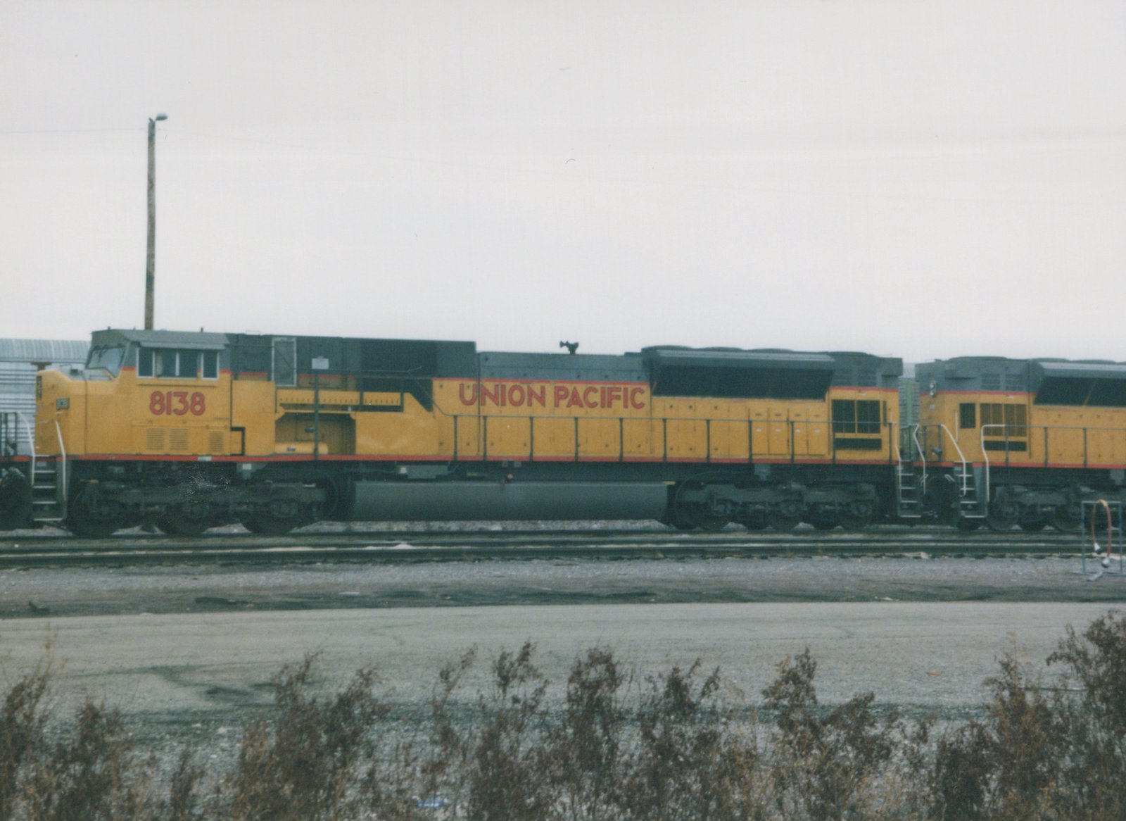 A yellow and gray Union Pacific locomotive with the number 8138 on its side is stopped on the tracks in a rail yard, with another locomotive visible behind it under a gray sky.