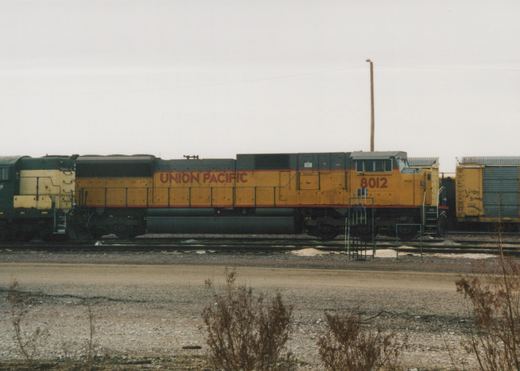 A side view of a yellow Union Pacific diesel locomotive, number 8012, sitting on tracks in a gravel yard with a boxcar in the background.