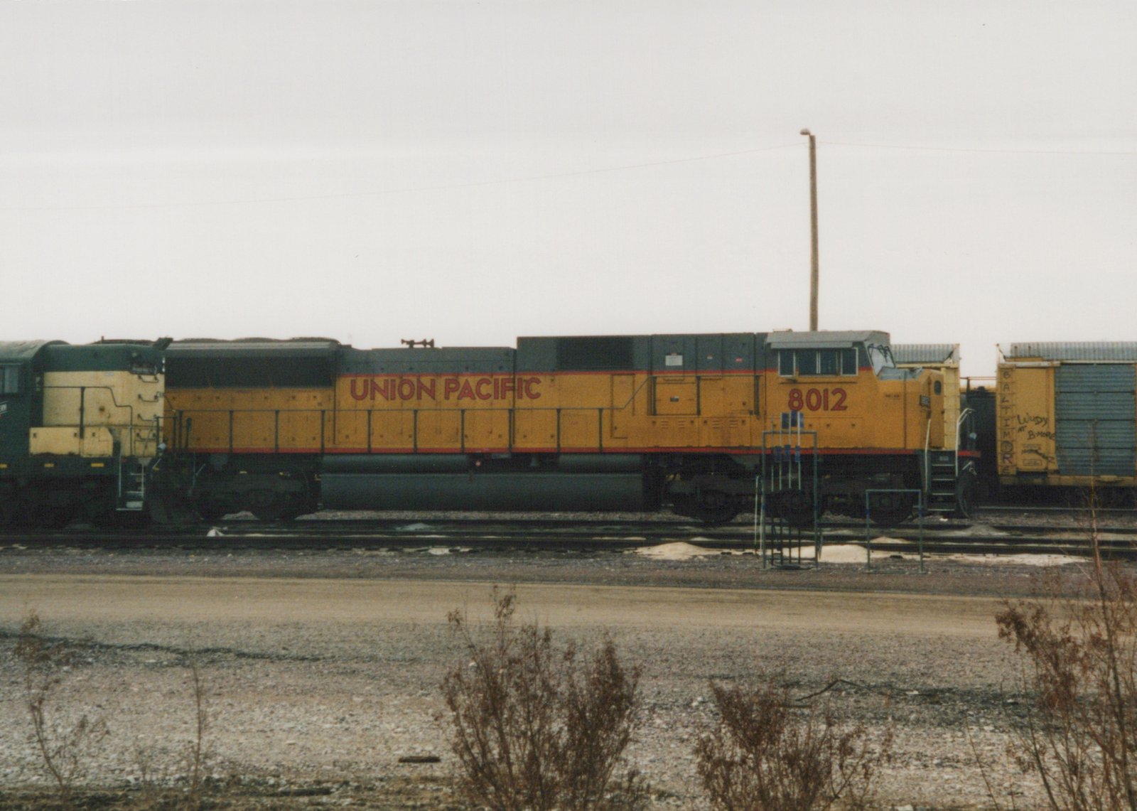 A side view of a yellow Union Pacific diesel locomotive, number 8012, sitting on tracks in a gravel yard with a boxcar in the background.