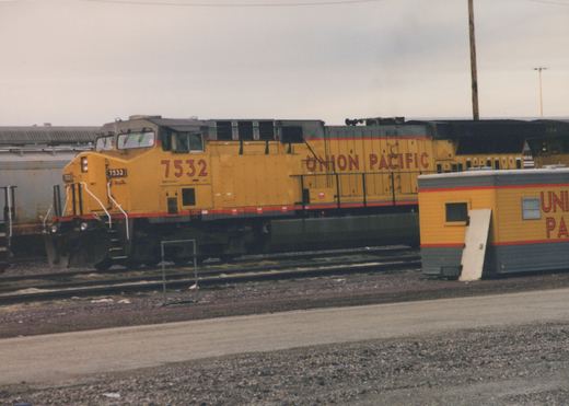 A yellow Union Pacific diesel locomotive, number 7532, is parked on railroad tracks in a yard, with a small shed and other railcars visible in the background.