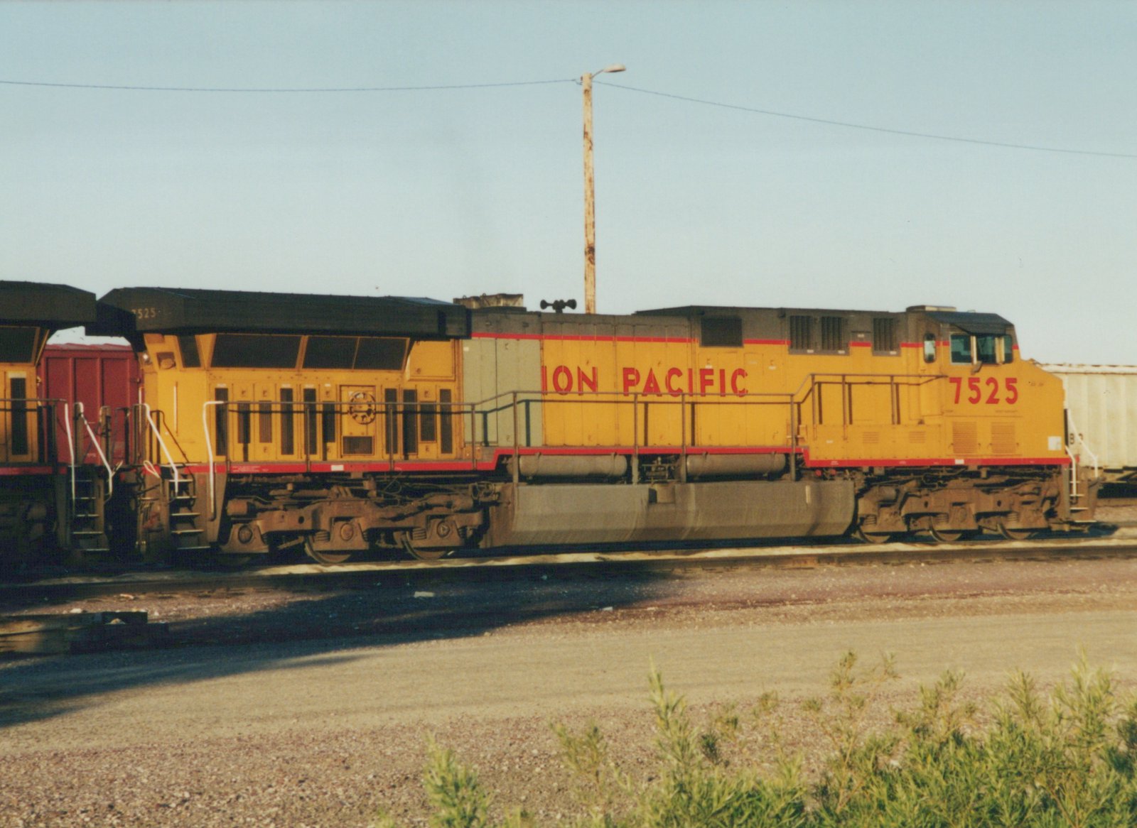 A yellow, gray, and red Union Pacific locomotive numbered 7525 is parked on a railroad track, with a red freight car attached to its front and a light-colored freight car behind it.