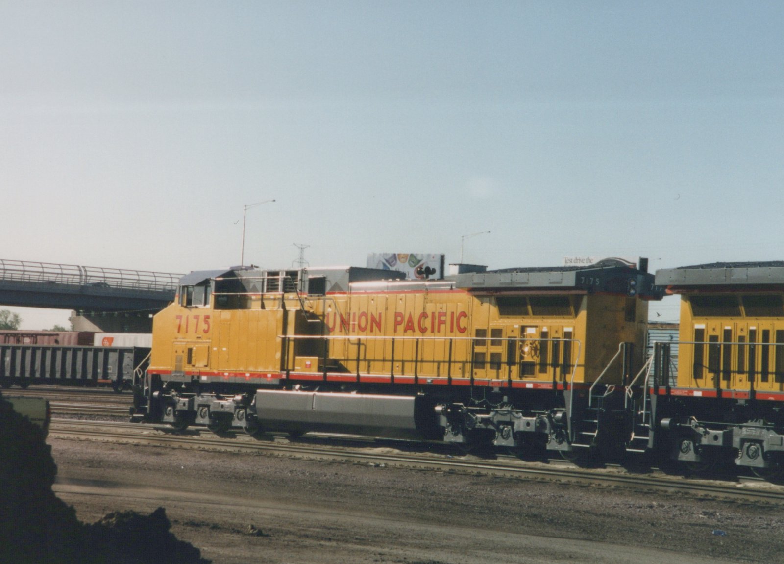 A yellow and red Union Pacific diesel locomotive, number 7175, is shown on a set of tracks with a bridge and freight cars in the background.
