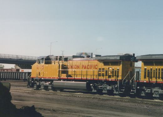 A yellow and red Union Pacific diesel locomotive, number 7175, is shown on a set of tracks with a bridge and freight cars in the background.
