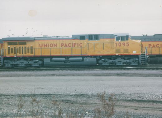 Side view of a yellow and gray Union Pacific diesel locomotive numbered 7095, standing on tracks in a gravel yard with bare ground and overcast sky.