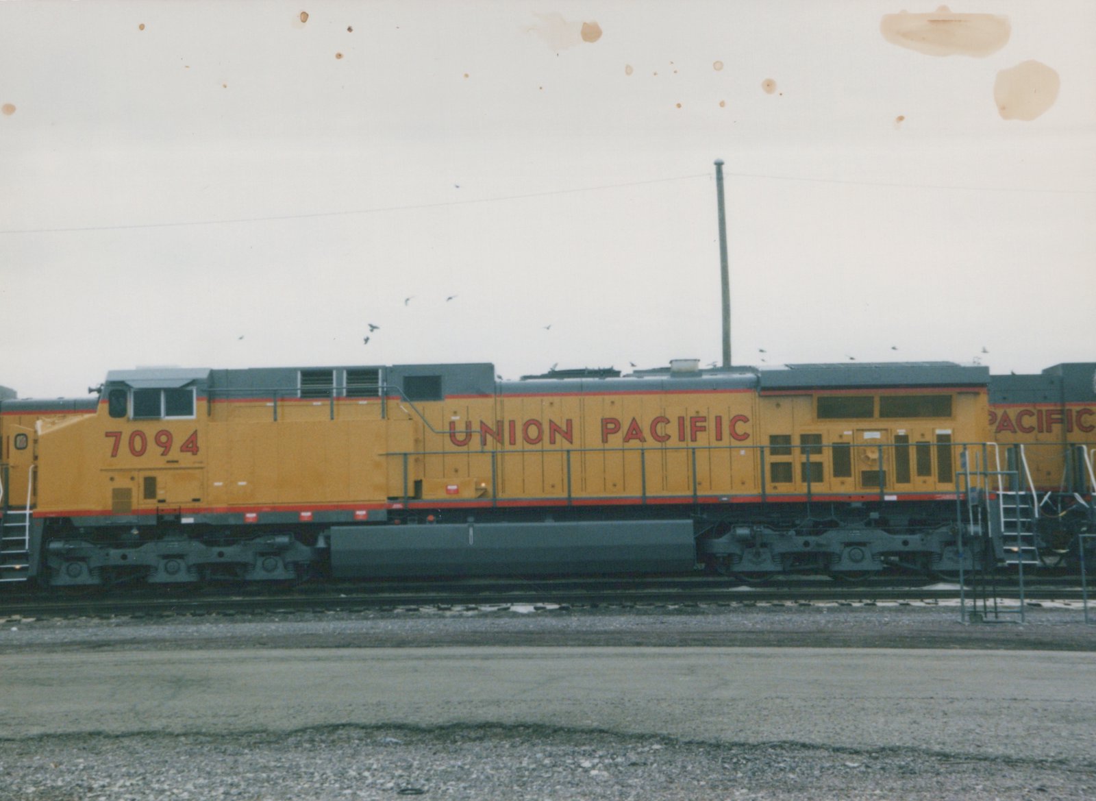 Side view of a yellow and gray Union Pacific diesel locomotive, number 7094, parked on tracks in a rail yard under a gray sky.