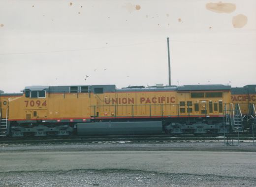 Side view of a yellow and gray Union Pacific diesel locomotive, number 7094, parked on tracks in a rail yard under a gray sky.