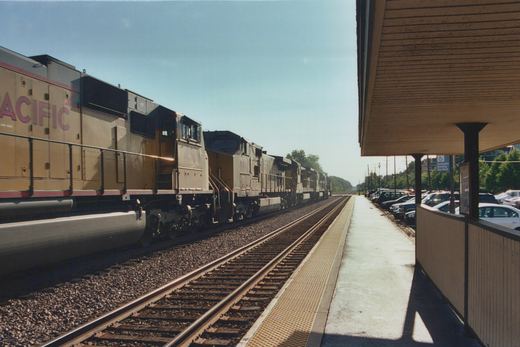 A long line of Santa Fe Railway diesel locomotives, including number 4[?]00 and 9014, are parked on tracks beside a station platform with a covered waiting area and a row of parked cars beyond it.