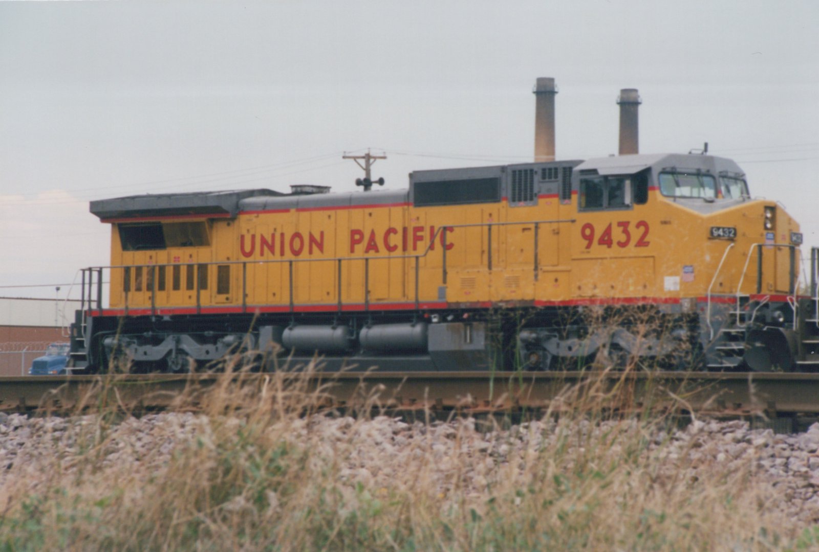 A yellow Union Pacific diesel locomotive numbered 9432 sits on railroad tracks, with overgrown grass and a fence in the foreground.