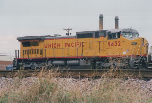 A yellow Union Pacific diesel locomotive numbered 9432 sits on railroad tracks, with overgrown grass and a fence in the foreground.