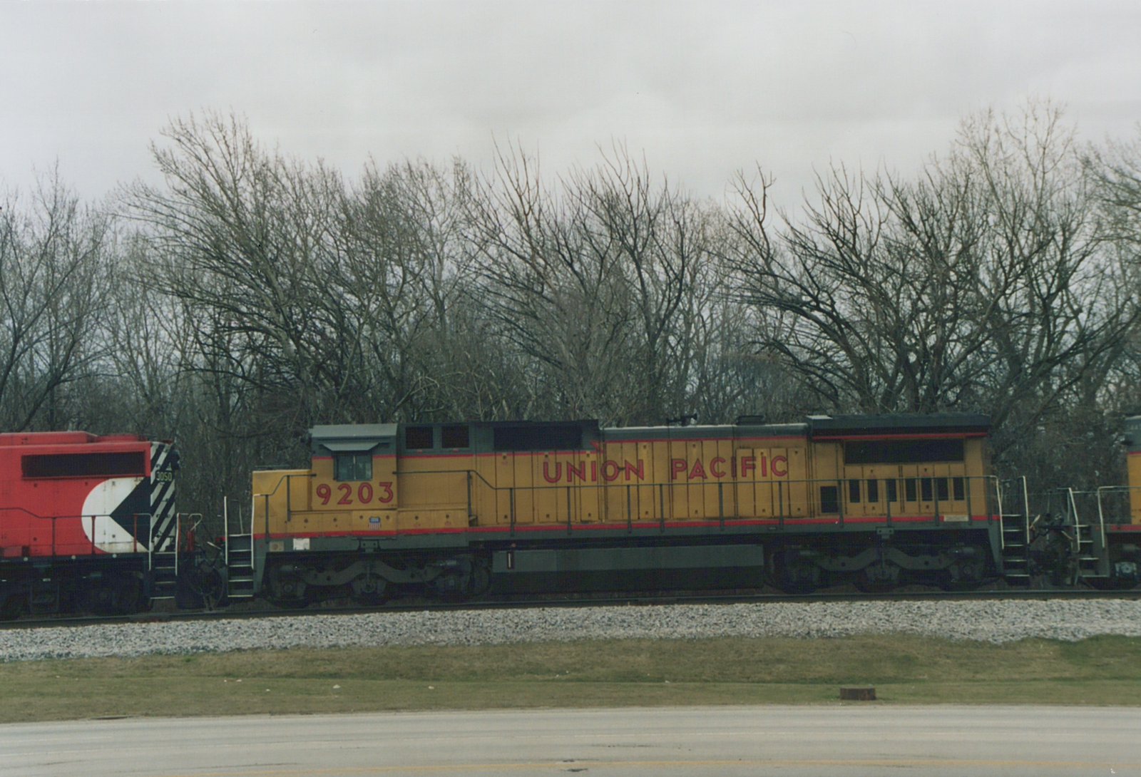 Side view of two diesel locomotives, one yellow Union Pacific and one red Chicago and North Western, on parallel tracks with bare trees in the background under a gray sky.
