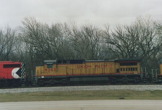 Side view of two diesel locomotives, one yellow Union Pacific and one red Chicago and North Western, on parallel tracks with bare trees in the background under a gray sky.