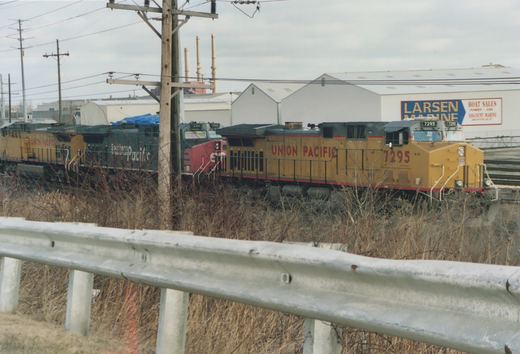 A yellow Union Pacific locomotive numbered 7295 leads a train past a guardrail, with a red and gray Southern Pacific locomotive behind it, in front of a building that says Larsen Marine.