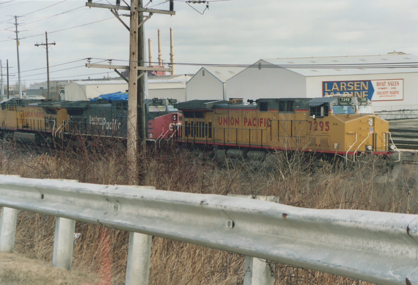 A yellow Union Pacific locomotive numbered 7295 leads a train past a guardrail, with a red and gray Southern Pacific locomotive behind it, in front of a building that says Larsen Marine.