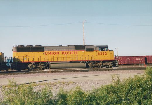 A yellow Union Pacific locomotive, number 6282, stands on railroad tracks in a yard next to maroon Wisconsin Central boxcars under a clear blue sky with green bushes in the foreground.