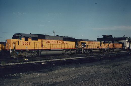 Three yellow and gray Union Pacific diesel locomotives, numbered 5036, 105, and 5003, sit side by side on multiple tracks in a railroad yard under a blue sky.
