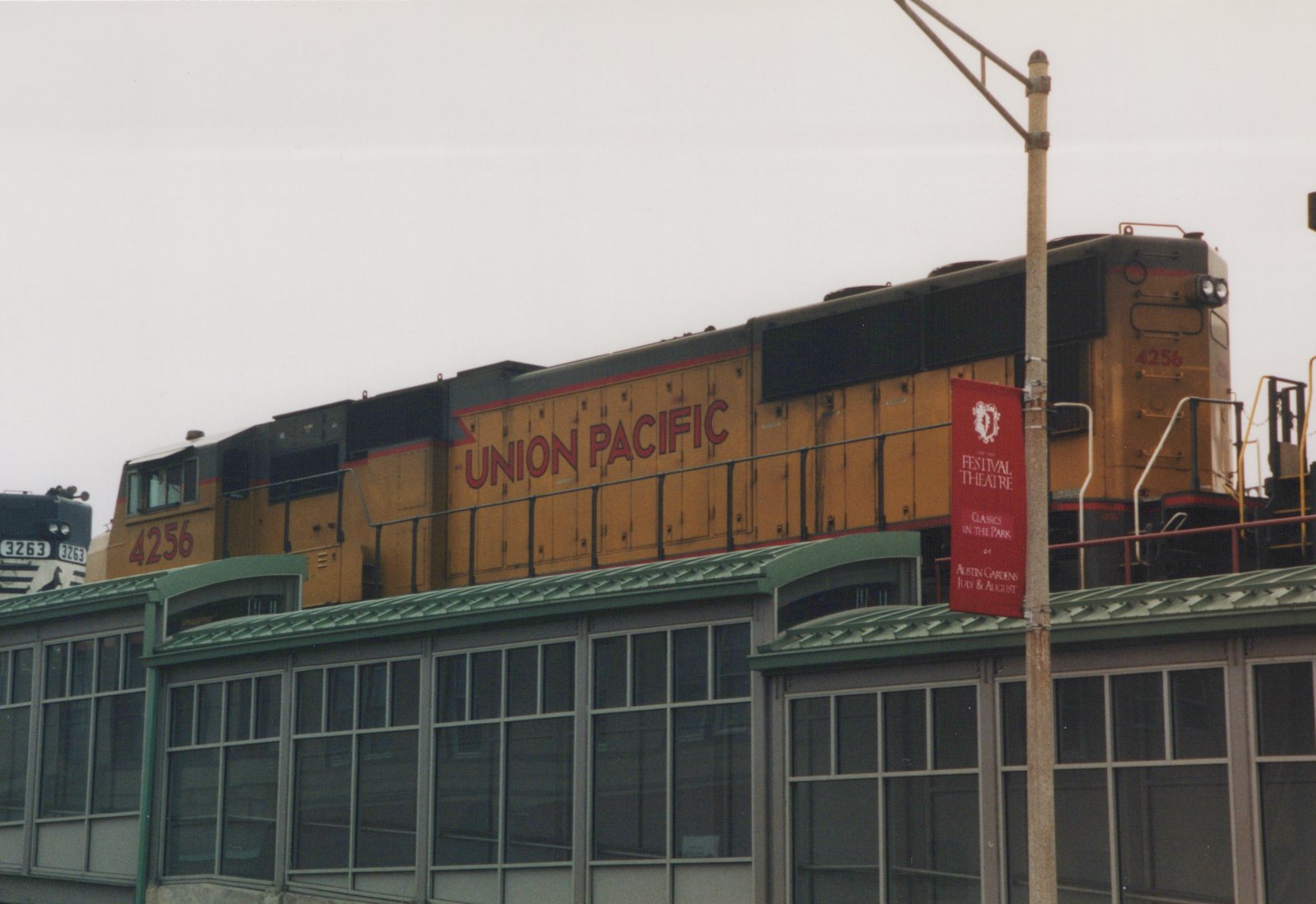 A yellow and black Union Pacific diesel locomotive numbered 4256 travels on an elevated track above a station platform, with a red banner for the Festival Theatre hanging on a nearby pole.